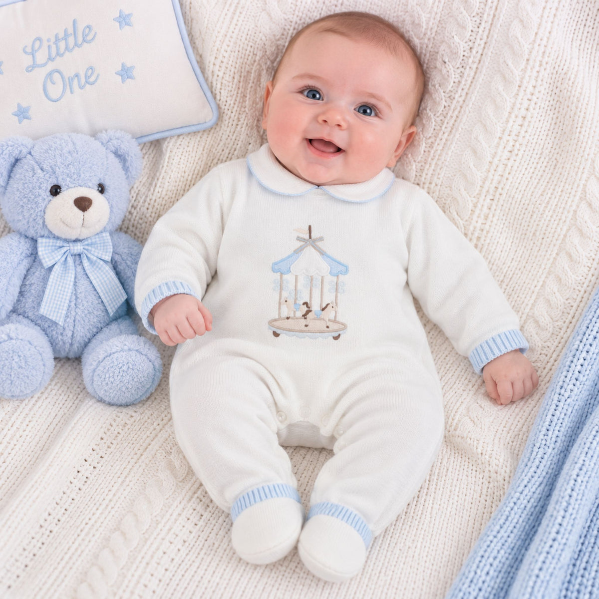 A baby lying on a cream cable-knit blanket next to a blue teddy bear and a pillow that says 'Little One'. The baby is wearing an off-white footed sleeper with light blue ribbed cuffs and hem, and a carousel graphic on the chest.