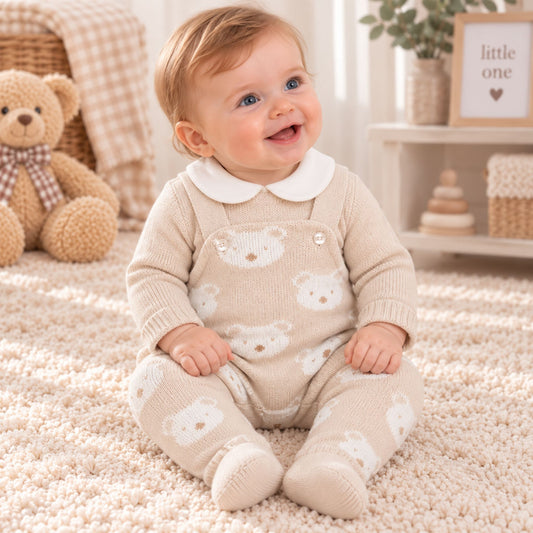 A baby sitting on a cream shag rug wearing a beige knit footed romper with a white Peter Pan collar and white teddy bear face patterns. The background shows a cozy nursery setting with a teddy bear, baskets, and a shelf.