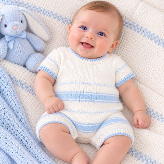 A smiling baby lying on a cream knit blanket with light blue stripes, wearing a matching cream and light blue striped short-sleeve romper. A light blue plush bunny toy sits beside the baby.