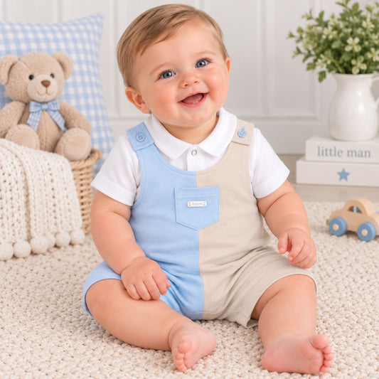 A smiling baby sitting on a cream carpet wearing a color-blocked overall romper with light blue and beige panels over a white short-sleeve shirt. The background features a teddy bear, a blue-and-white check pillow, a white vase with greenery, and a small wooden toy car.