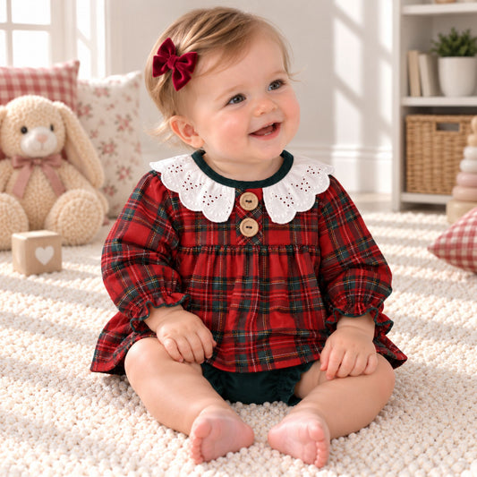 A baby girl sitting on a white textured rug in a cozy room, wearing a red and green plaid long-sleeve top with a white lace Peter Pan collar and two wooden buttons.