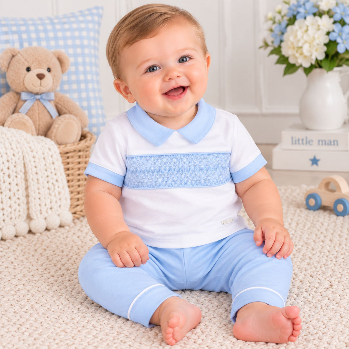 A smiling baby sitting on a cream carpet wearing a white short-sleeve polo shirt with a light blue collar and smocked chest panel, paired with light blue pants. The background features a teddy bear, a blue gingham pillow, a white vase of flowers, and a small wooden toy car.