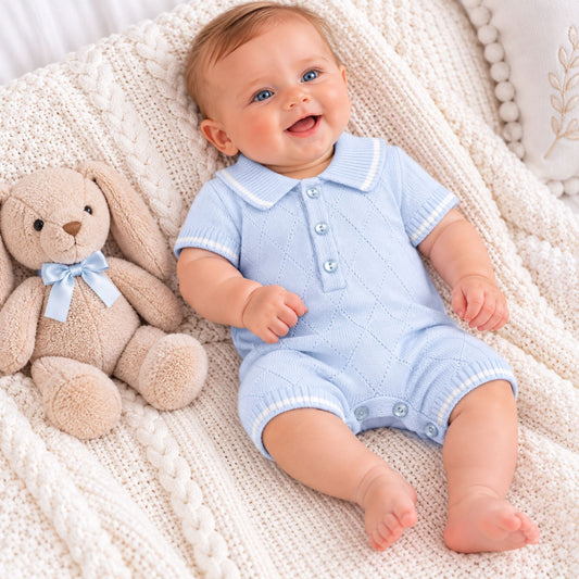 A smiling baby wearing a light blue short-sleeve knit romper lies on a cream cable-knit blanket next to a beige stuffed bunny with a light blue bow.