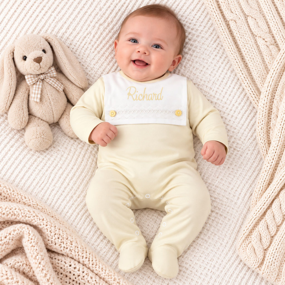 A smiling baby lying on a cream knit blanket next to a plush bunny, wearing a pale yellow footed sleeper with a white bib panel personalized with the name 'Richard' in gold embroidery.