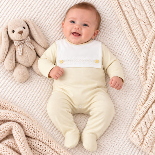 A smiling baby lying on a cream knit blanket next to a plush bunny, wearing a pale yellow footed sleeper with a white bib panel personalized with the name 'Richard' in gold embroidery.
