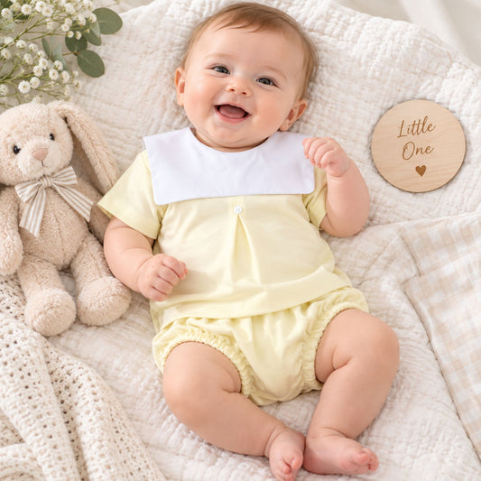 A smiling baby lying on a cream textured blanket, wearing a pale yellow short-sleeve romper with a contrasting white collar. A plush cream bunny toy sits to the left, and a round wooden sign reading 'Little One' is to the right.