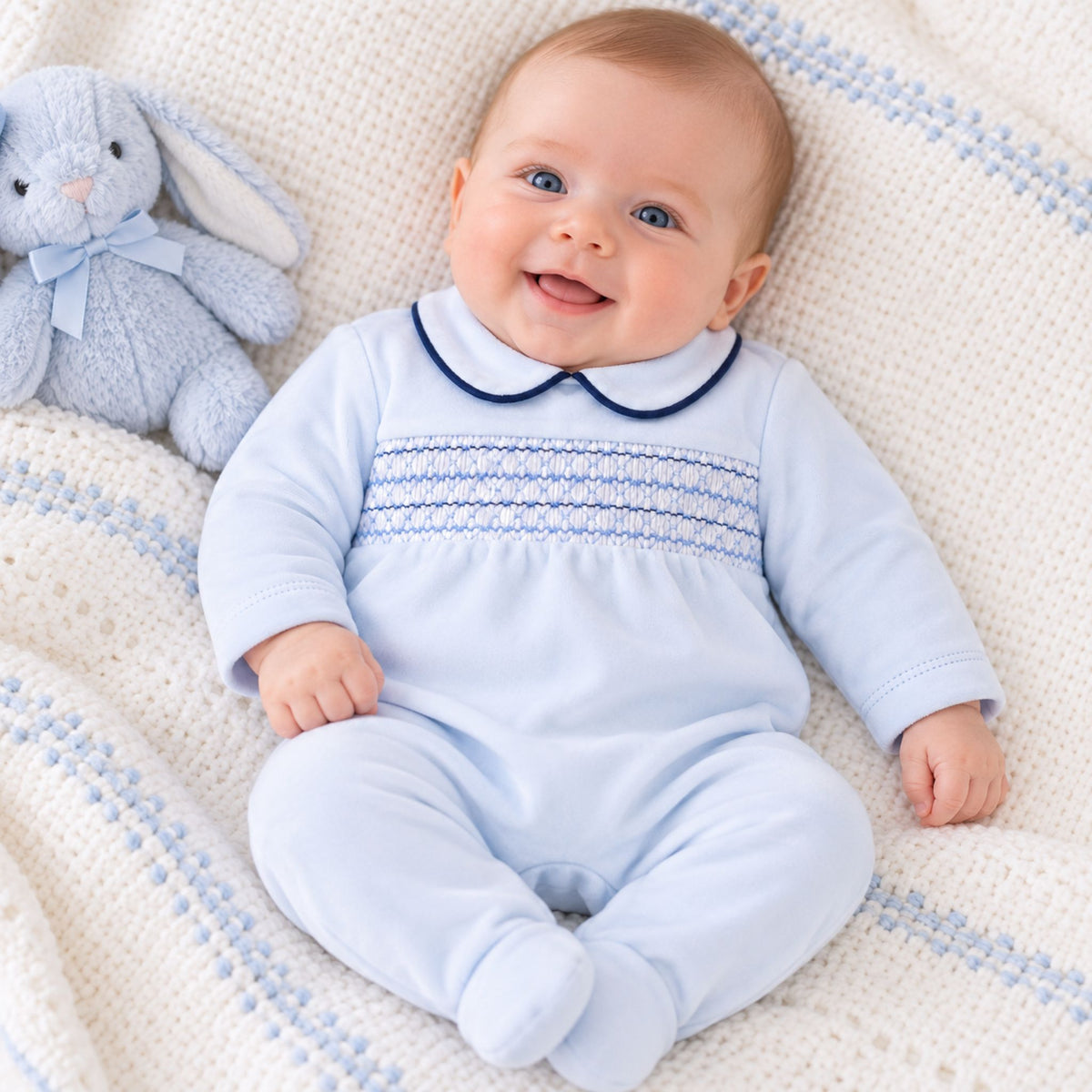 A smiling baby wearing a light blue footed onesie with a dark blue Peter Pan collar and smocked chest details, lying on a cream knit blanket with a light blue bunny plush toy next to them.