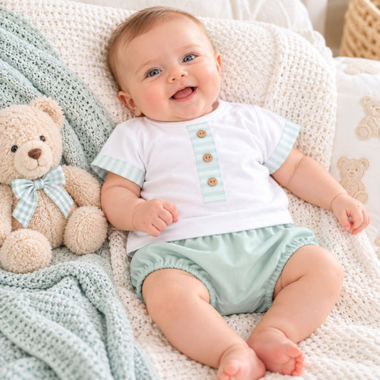 A smiling baby lying on a white knit blanket next to a beige teddy bear, wearing a white short-sleeve top with pale green striped cuffs and a vertical row of three wooden buttons, paired with pale green shorts.