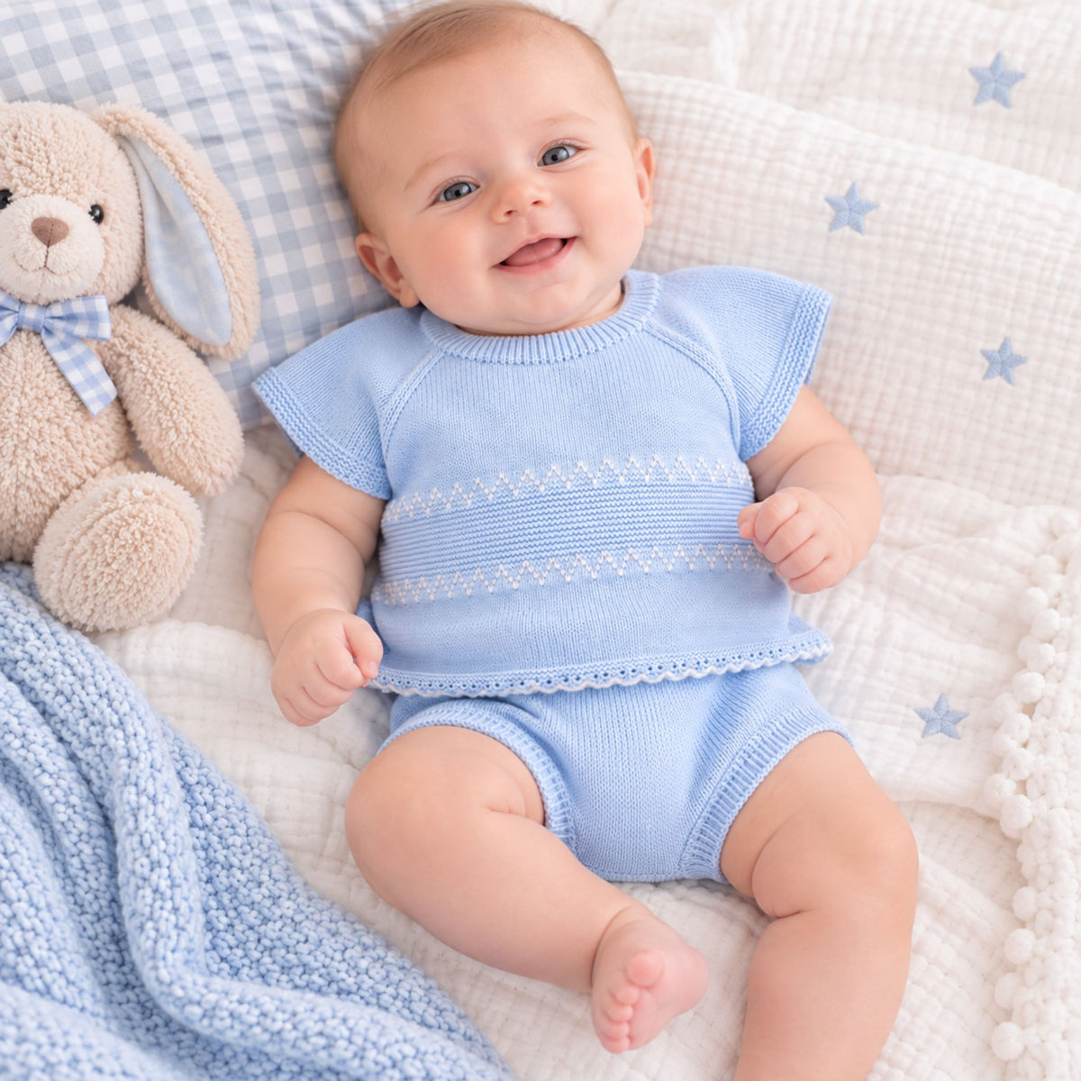 A smiling baby lying on a white textured blanket with blue star prints, wearing a light blue knit short-sleeve top and matching bloomers. A beige plush bunny toy and a blue gingham pillow are beside the baby.