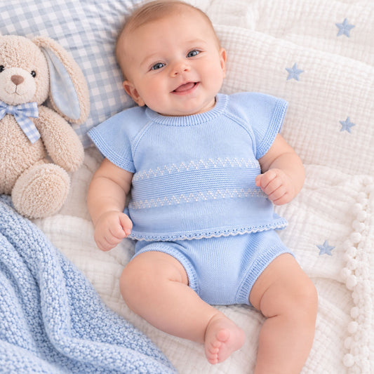 A smiling baby lying on a white textured blanket with blue star prints, wearing a light blue knit short-sleeve top and matching bloomers. A beige plush bunny toy and a blue gingham pillow are beside the baby.