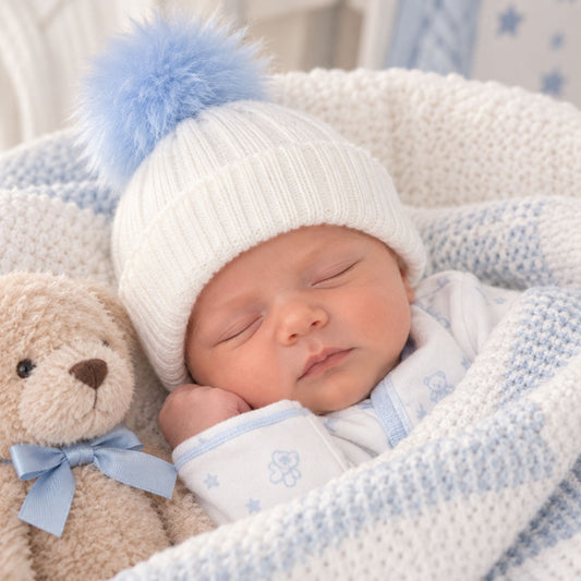 A sleeping newborn baby wearing a white knit beanie with a fluffy light blue pom-pom, nestled in a white and light blue blanket next to a beige teddy bear with a blue bow.