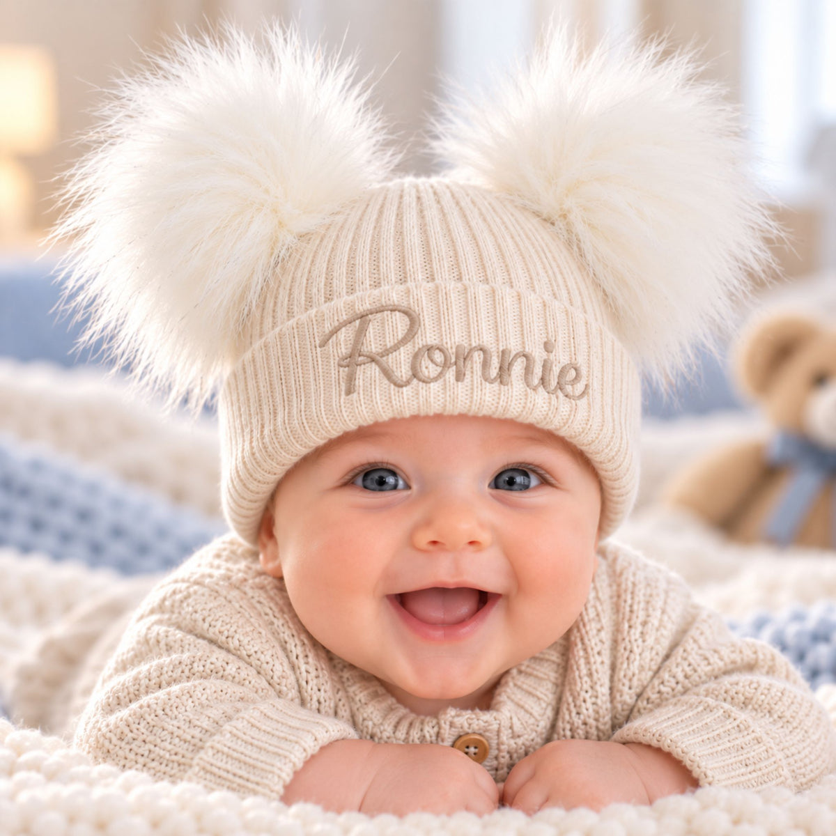 A smiling baby wearing a beige ribbed knit pom-pom beanie with the name 'Ronnie' embroidered on the front, lying on a soft cream blanket.