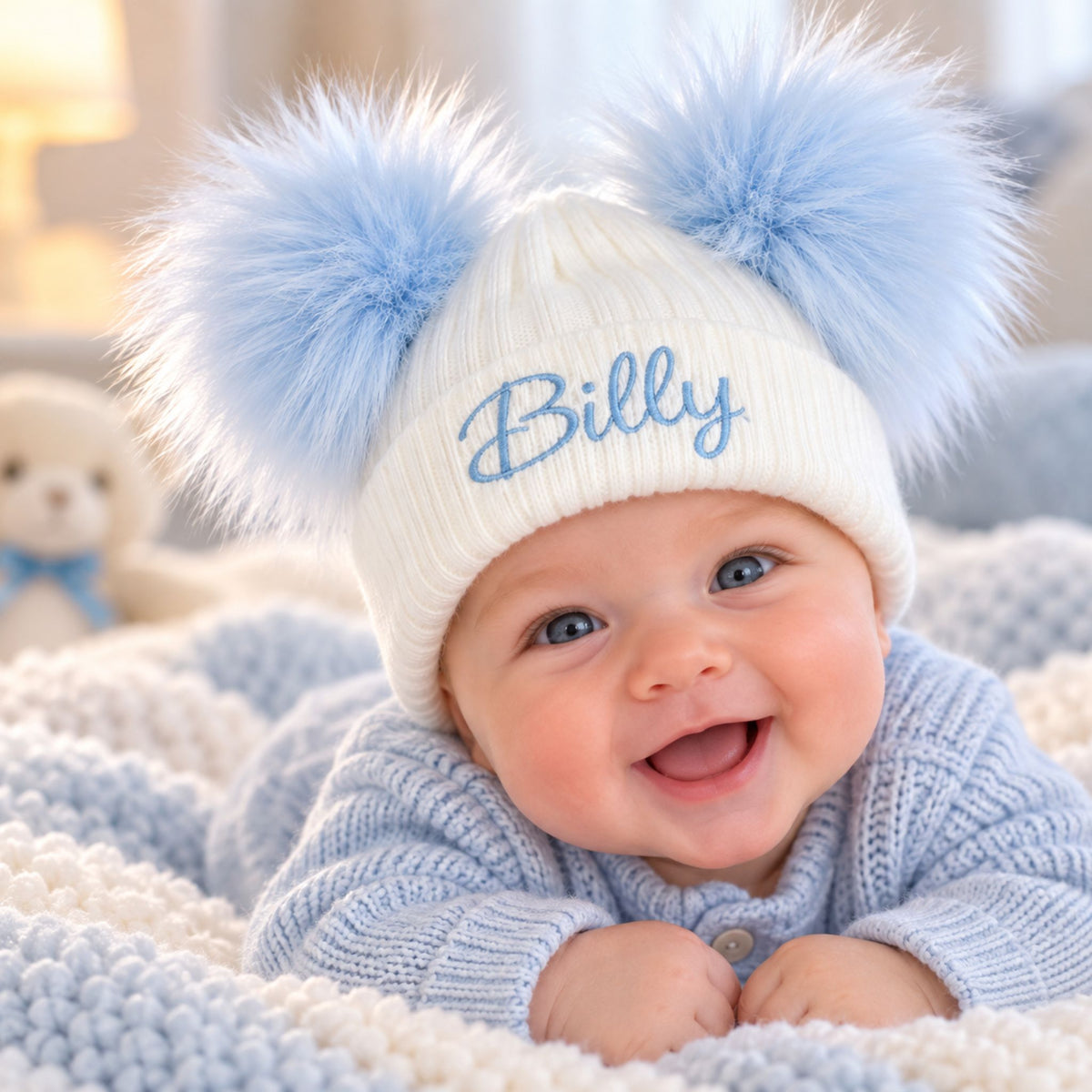 A smiling baby lying on a soft knit blanket, wearing a white  ribbed beanie with two fluffy light blue pompoms and the name 'Billy' embroidered in blue on the front.