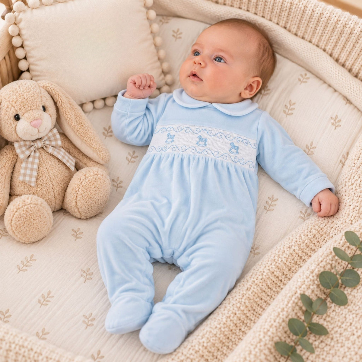A baby lying on a cream quilted bassinet wearing a light blue footed sleeper with embroidered details. A beige stuffed bunny with a checkered bow is sitting next to the baby.
