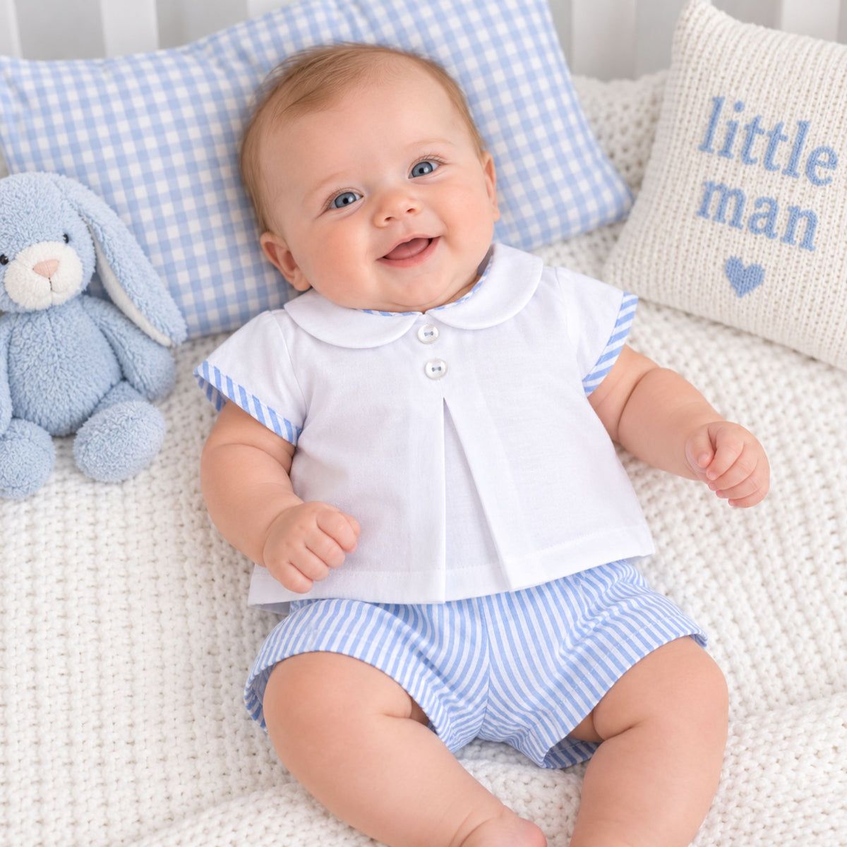 A smiling baby wearing a white short-sleeve button-front top with blue striped trim and matching blue-and-white striped shorts, sitting on a textured cream blanket with blue gingham pillows and a plush bunny.