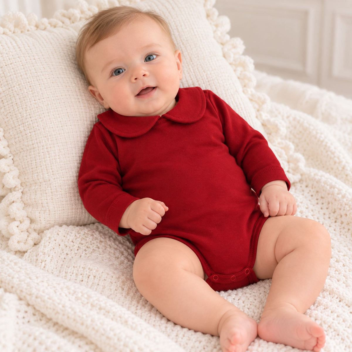 A baby wearing a long-sleeve red romper with a Peter Pan collar, sitting on a cream knit blanket and pillow.
