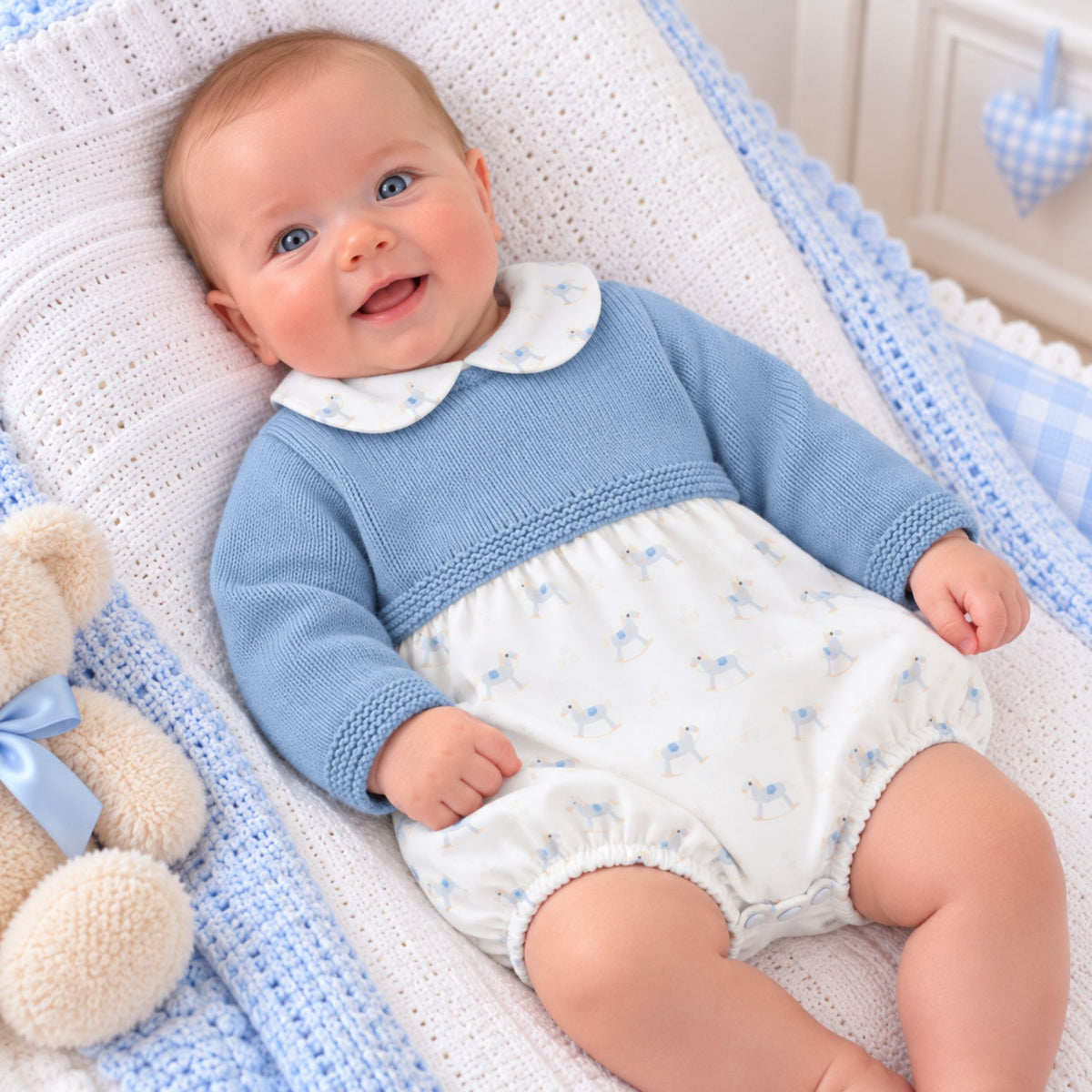 A smiling baby lying on a white and blue crocheted bassinet or cushion, wearing a light blue knit romper with a white Peter Pan collar and small blue floral print on the lower half. A beige teddy bear is partially visible at the left edge.