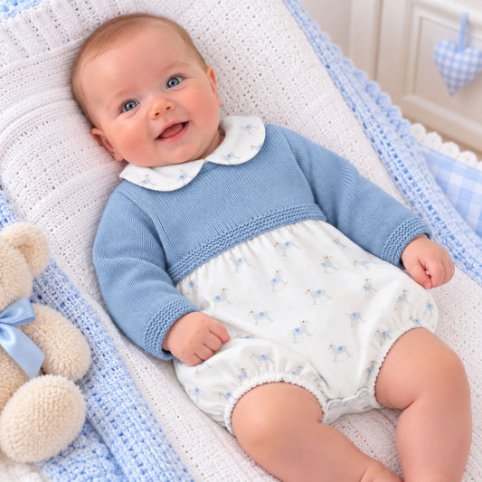 A smiling baby lying on a white and blue crocheted bassinet or cushion, wearing a light blue knit romper with a white Peter Pan collar and small blue floral print on the lower half. A beige teddy bear is partially visible at the left edge.