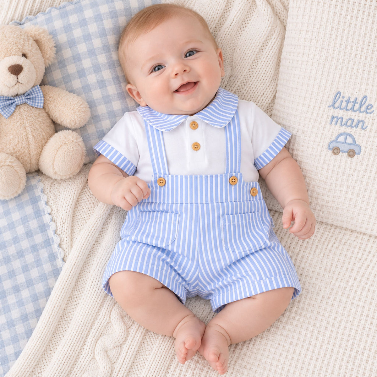 A smiling baby lying on a cream knit blanket with a blue gingham pillow and a teddy bear, wearing a white short-sleeve top with blue striped collar and sleeves, plus blue-and-white striped overalls with gold buttons. A pillow reads 'little man'.