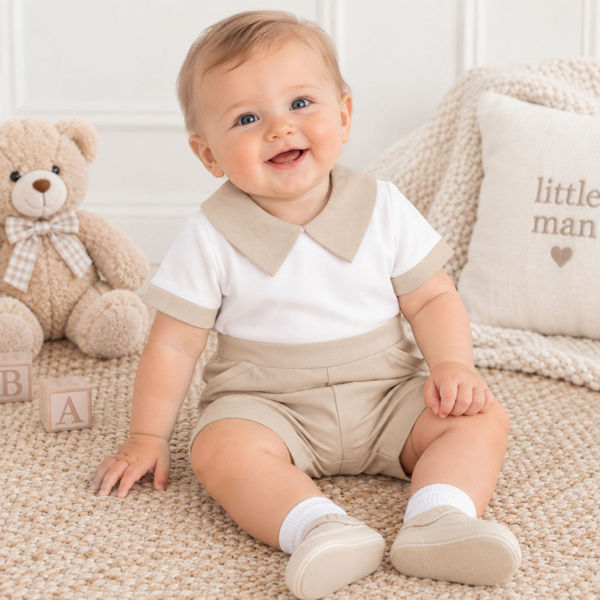 A baby sitting on a beige woven rug wearing a white short-sleeve polo shirt with a beige collar and matching beige shorts. The baby is surrounded by a plush teddy bear, wooden blocks, and a knitted blanket with a pillow.