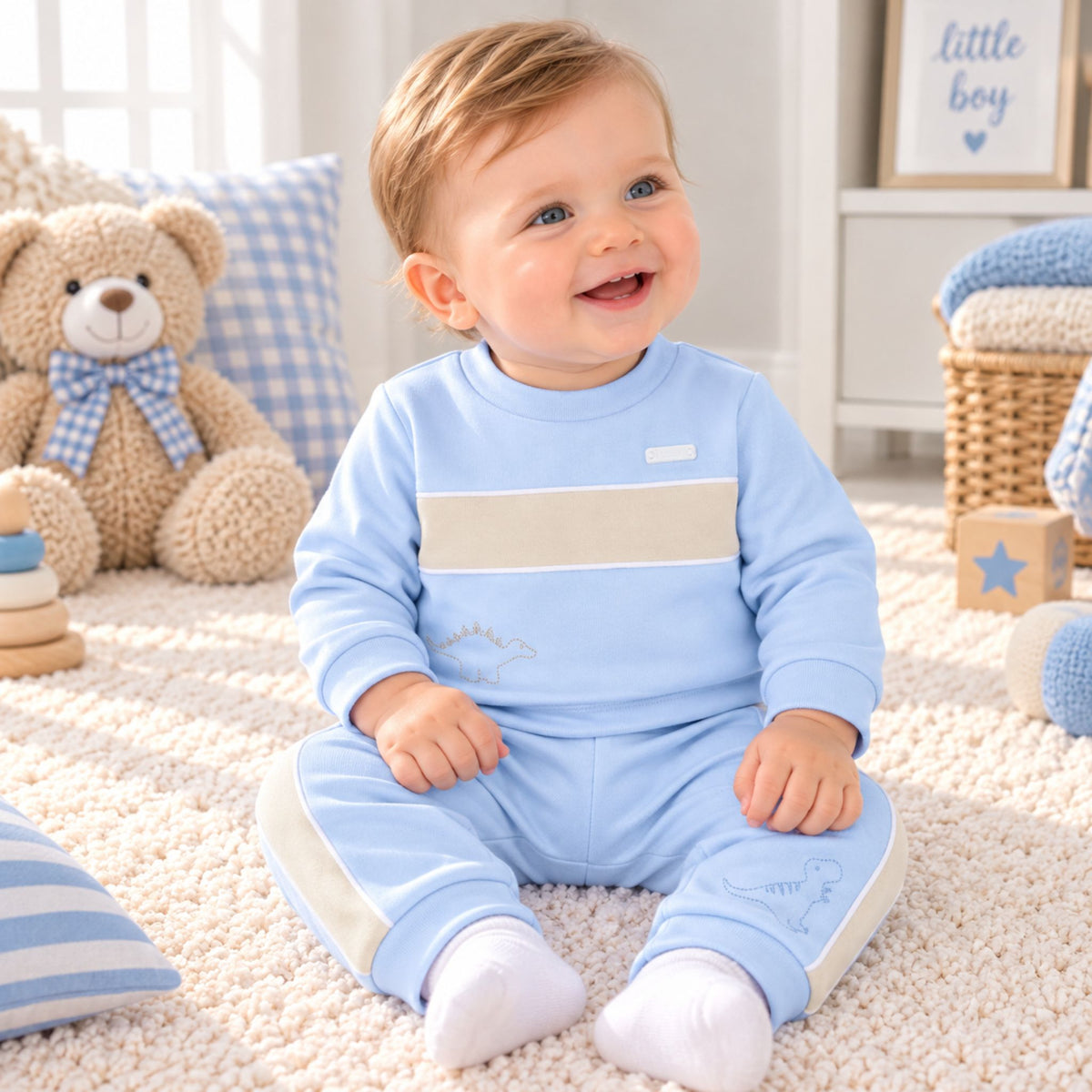 A baby sitting on a cream carpet in a cozy nursery, wearing a light blue two-piece outfit with a beige chest stripe and matching pants. The background features a teddy bear, blue gingham pillows, and a basket.