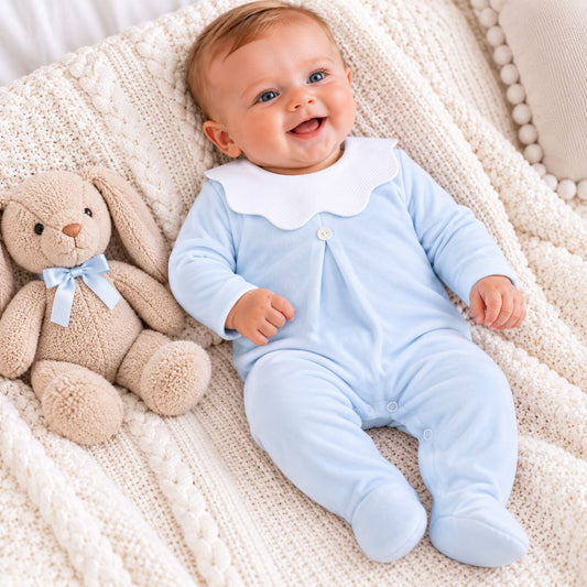 A baby lying on a cream knit blanket next to a plush teddy bear, wearing a light blue footed sleeper with a white Peter Pan collar.