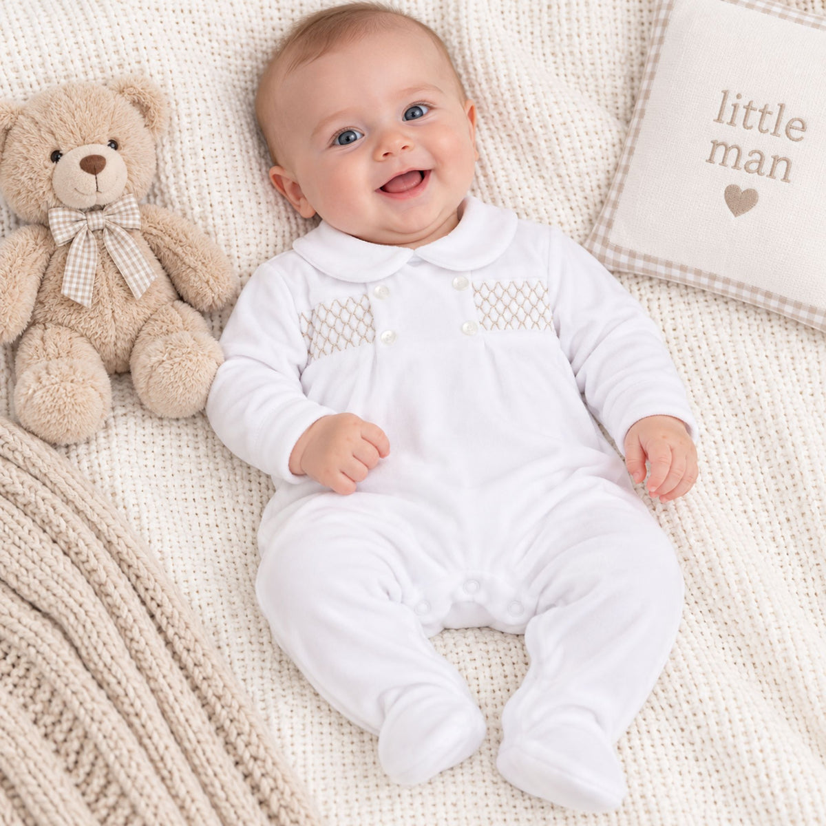 A smiling baby lying on a cream knit blanket next to a teddy bear and a pillow that says 'little man', wearing a white footed onesie with decorative knit detailing on the chest.