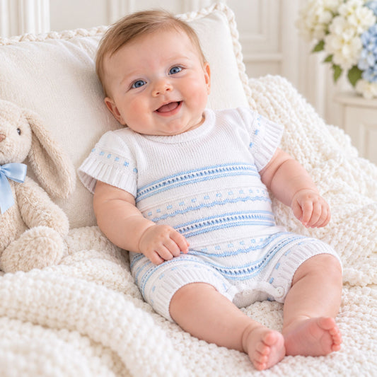 A smiling baby sitting on a cream knit blanket with a white and light blue patterned romper, posed next to a plush bunny toy.