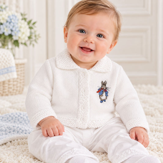 A smiling baby sitting on a cream rug wearing a white knit cardigan with a Rabbit embroidered motif on the chest, paired with white pants.