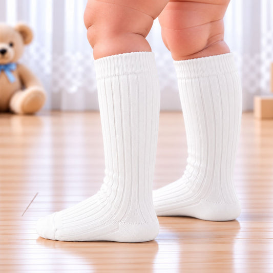 White ribbed knee-high socks worn on a child's legs, standing on a light wooden floor with a soft-focus teddy bear in the background.