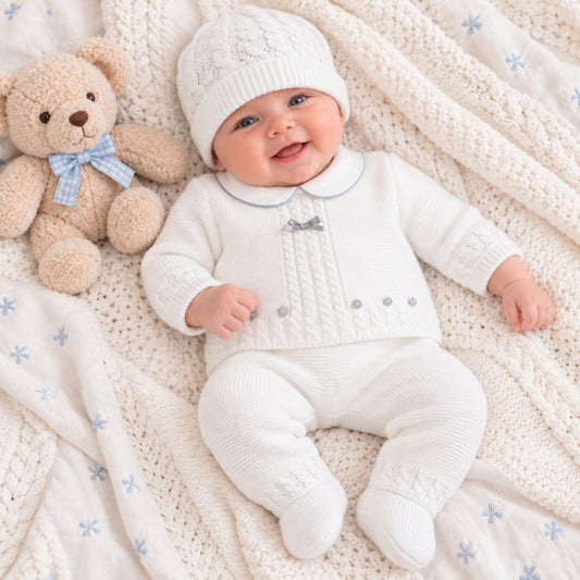 A smiling baby lying on a cream cable-knit blanket with a teddy bear, wearing a matching cream knit hat, cardigan, and footed pants set with small blue accents.