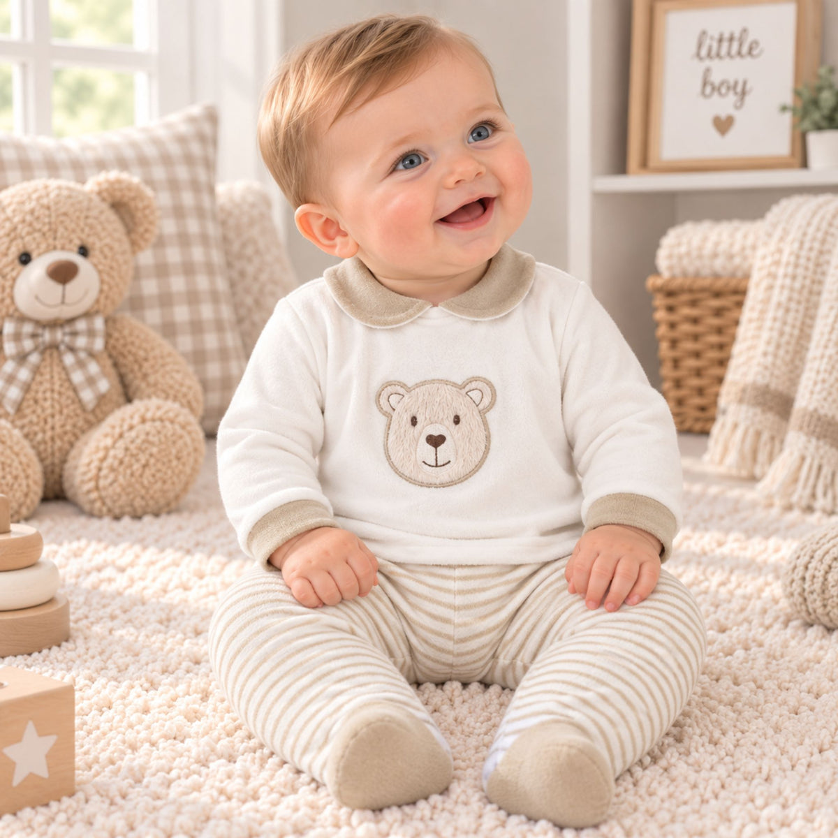 A smiling baby sitting on a cream rug wearing a cream long-sleeve top with a teddy bear face and matching striped pants. The scene is styled with a plush teddy bear, pillows, and a basket in a bright, cozy room.