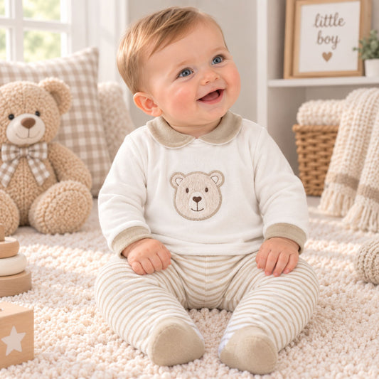 A smiling baby sitting on a cream rug wearing a cream long-sleeve top with a teddy bear face and matching striped pants. The scene is styled with a plush teddy bear, pillows, and a basket in a bright, cozy room.
