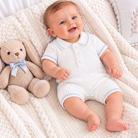 A baby lying on a cream cable-knit blanket next to a beige stuffed bunny, wearing a white short-sleeve polo romper with light blue trim.