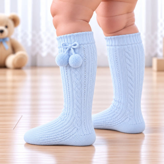 A close-up of a child's legs wearing light blue cable-knit socks with pom-pom details, standing on a wooden floor with a soft-focus teddy bear in the background.