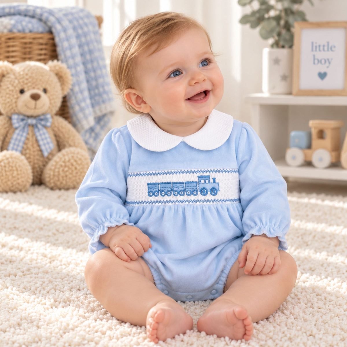 A smiling baby sitting on a cream carpet in a nursery, wearing a light blue long-sleeve romper with a white Peter Pan collar and a blue embroidered train motif across the chest.