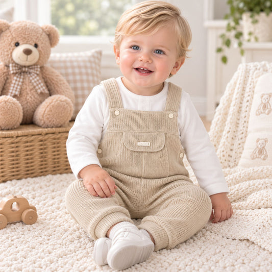 A smiling toddler sits on a cream knit rug in a bright nursery, wearing beige corduroy overall-style pants over a white long-sleeve top. A teddy bear sits on a wicker basket in the background, with a soft blanket and toys around them.