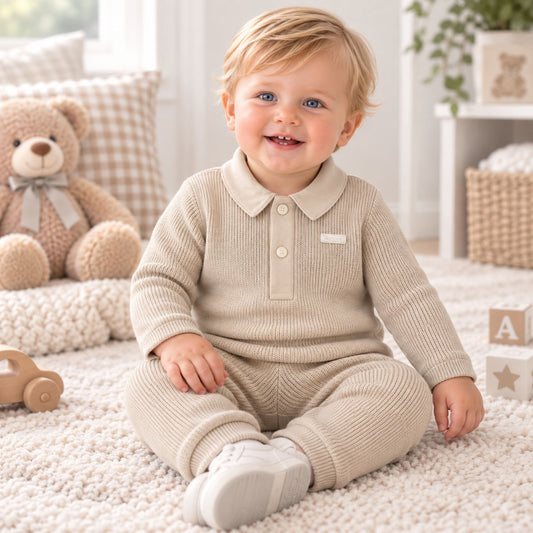 A smiling toddler sitting on a cream carpet in a bright nursery, wearing a ribbed knit long-sleeve polo romper in a light oatmeal beige color. The outfit includes matching pants and white shoes.