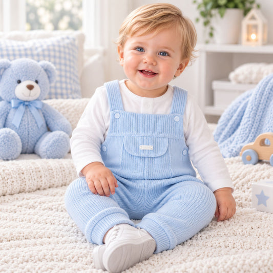 A smiling toddler sitting on a cream knit rug in a bright nursery, wearing light blue ribbed knit overall over a white long-sleeve shirt. A blue teddy bear, pillows, and soft nursery decor are visible in the background.