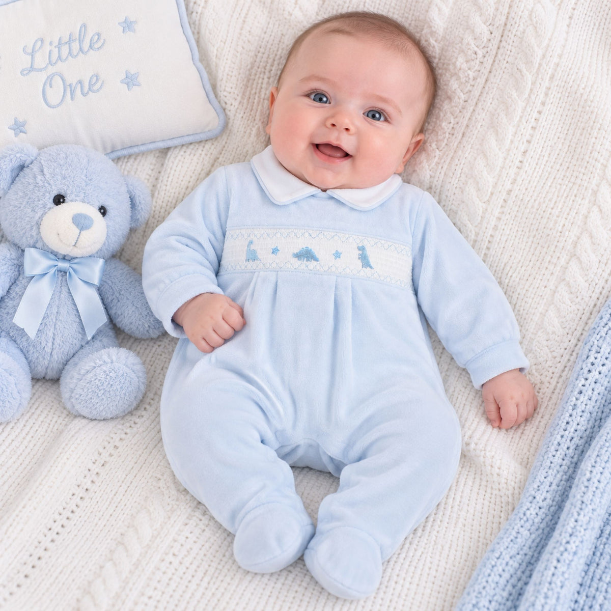 A baby lying on a cream cable-knit blanket, wearing a light blue footed onesie with a white Peter Pan collar and a decorative chest panel. A plush blue teddy bear and a white pillow with blue embroidery are beside the baby.