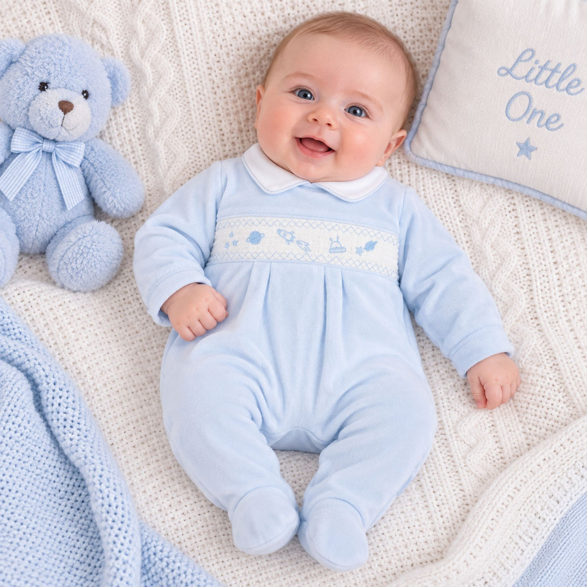 A baby lying on a cream knit blanket wearing a light blue footed romper with a white collar and a decorative embroidered chest panel. A blue teddy bear sits to the left, and a cream pillow with blue lettering is to the right.