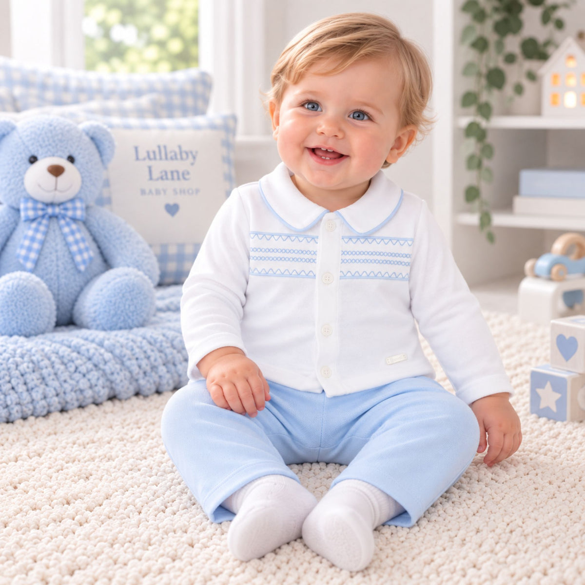 A baby sitting on a cream carpet in a bright nursery, wearing a white long-sleeve button-front top with light blue embroidered trim and light blue pants. A blue teddy bear and a pillow reading 'Lullaby Lane Baby Shop' are visible in the background.