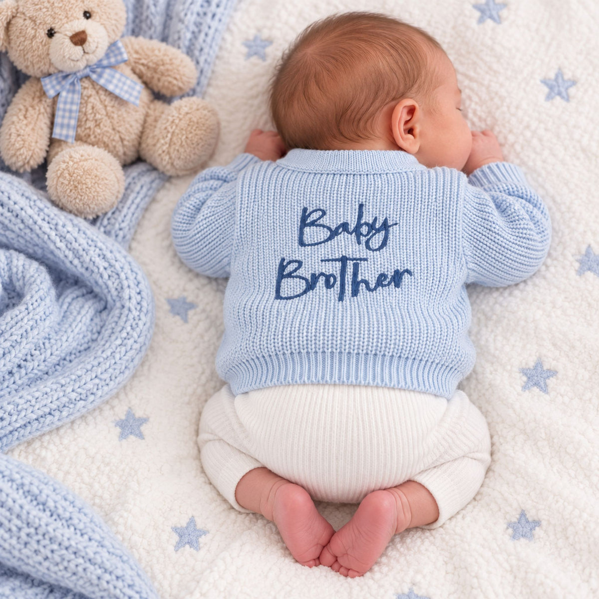 A baby lying on their side on a white fleece blanket with blue star prints, wearing a light blue ribbed knit cardigan with 'Baby Brother' embroidered in navy blue on the back, and white ribbed bodysuit bottoms. A teddy bear and a blue knit blanket are visible next to the baby.
