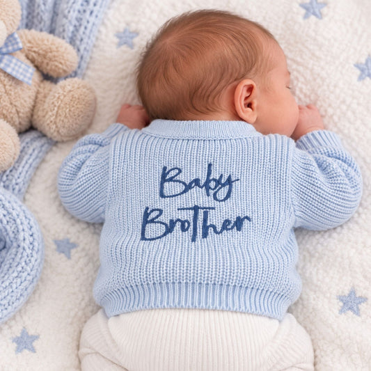 A baby lying on their side on a white fleece blanket with blue star prints, wearing a light blue ribbed knit cardigan with 'Baby Brother' embroidered in navy blue on the back, and white ribbed bodysuit bottoms. A teddy bear and a blue knit blanket are visible next to the baby.