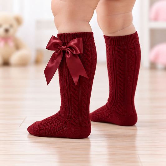 A close-up of a child's legs wearing deep red cable-knit knee-high socks with a large satin bow on one sock, standing on a light wood floor.