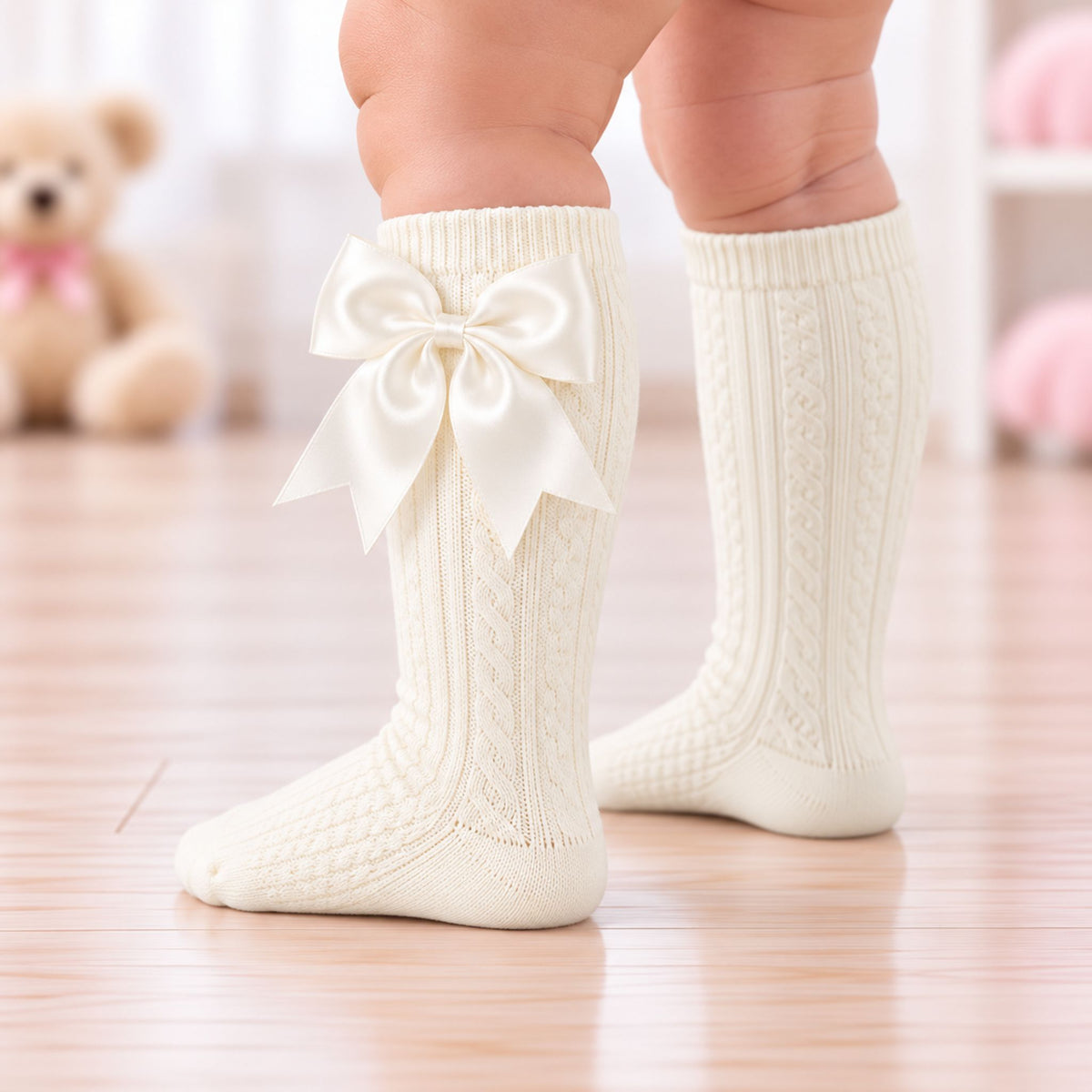 A close-up of a baby's legs wearing cream-colored, cable-knit knee-high socks with a large satin bow on the front of one sock, standing on a light wooden floor.