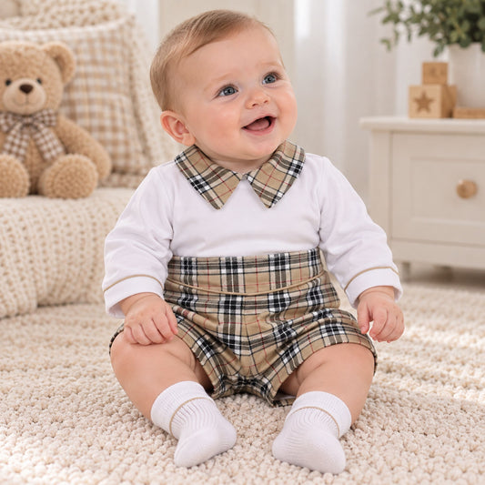 A baby sitting on a cream carpet wearing a white long-sleeve top with a tan and black plaid collar, matching plaid shorts, and white socks. A teddy bear and a white dresser are visible in the softly lit nursery background.