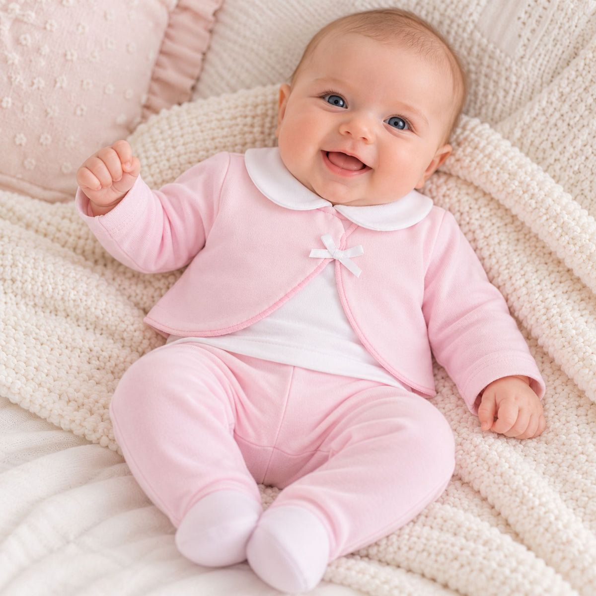 A smiling baby lying on a cream knit blanket, wearing a pink footed one-piece outfit with a white collar and a small bow detail.