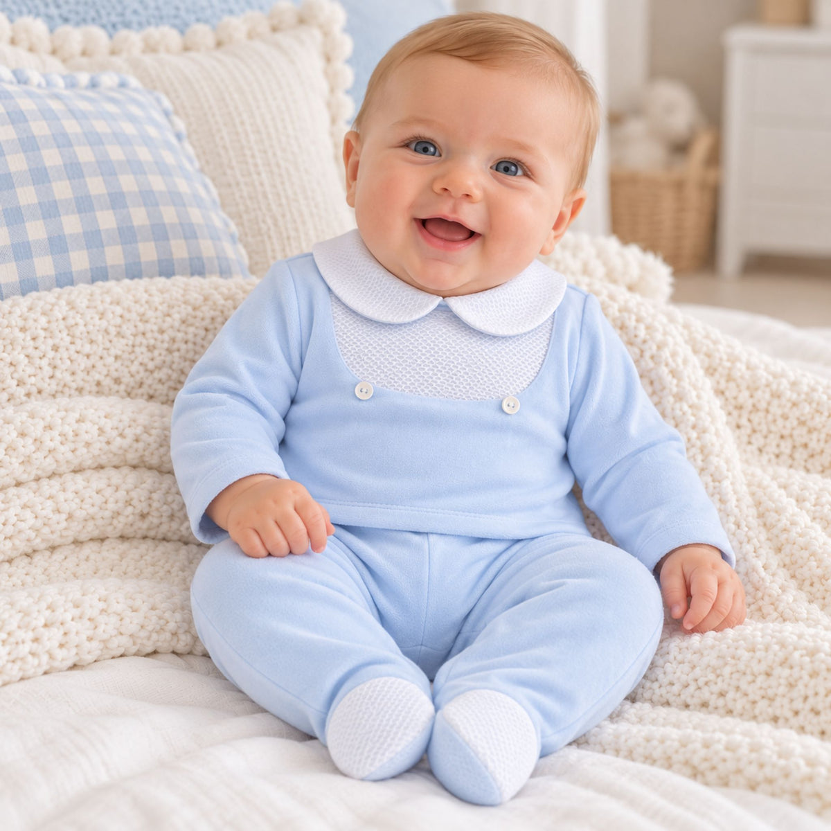 A smiling baby sitting on a cream knit blanket, wearing a light blue footed romper with a white Peter Pan collar and white button details.