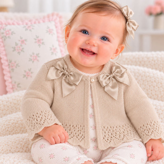 A smiling baby girl wearing a beige knitted cardigan with large satin bow details on the shoulders, layered over a white floral romper, sitting on a cozy cream blanket.